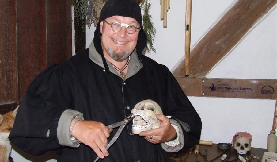 A man in a black Tudor smock and hat grinning humorously while holding a model of a skull and a pair of tooth pulling pliers