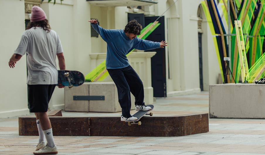 A skateboarder performs a trick on a skateable sculpture made of wood and metal on an urban street, showcasing skill and style amidst vibrant surround