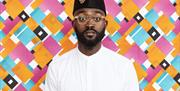 Inua stands against a brightly coloured, patterned backdrop, wearing glasses, a white shirt and a traditional hat.