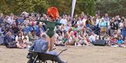 Two performers are outdoors in front of a large audience. One performer is dancing, wearing a bright green leotard and an oversized, colourful flower-