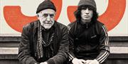 Two performers, one young and one older sat together in front of a large orange 55.