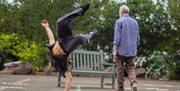 A performer doing breakdancing with an older performer walking past him to sit on a bench.