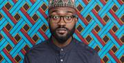 Inua stands against a brightly coloured, patterned backdrop, wearing glasses, a blue shirt and a traditional patterned hat.