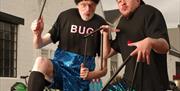 Two performers are outdoors wearing black hats, black T-shirts and sequin shorts. They have long black tubes attached to their fingers. The word "Bug"