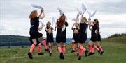 Eight Boss Morris dancers form a circle in a field, hopping as they fling white cloths into the air. They wear gold crowns, black tunics with sun and