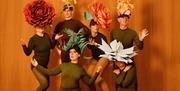 Six performers from a diverse range of ages, gender, shapes and sizes, pose as a flower arrangement against an orange-wood background. They are wearin