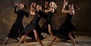 Four dancers in long black dresses raise their hands beside their faces, moving in front of a speckled brown background