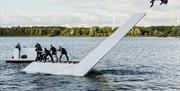 white staircase floating on water. One dancer is flipping backwards off the top of the staircase, about to land in the water.