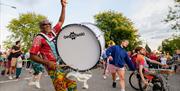 A musician in the street parade wearing a brightly coloured patterned outfit with a large drum strapped to his chest, surrounded by musicians and danc