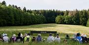 An audience sits in a field watching a large subtitled LED screen, with trees as a backdrop behind it.