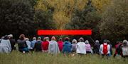 An audience sits on the grass watching a large red LED screen, with trees as a backdrop behind it.