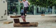 A young woman performs a trick on a skateboard on a skateable sculpture on an urban street, surrounded by greenery and onlookers.