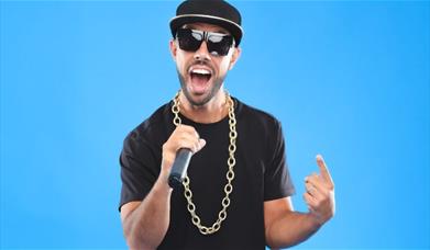 MC Grammar wearing a black hat and t-shirt holding a microphone against a blue background